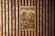 A Wyoming postage stamp featuring a dinosaur and a cowboy on horseback, mounted on a wooden wall.