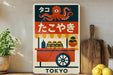 A colorful poster featuring an illustration of an ice cream vendor and an octopus, with the word "TOKYO" written on it. The poster is displayed on a wall next to a wooden cutting board and some kitchen utensils.
