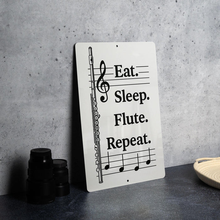 a sign with the text "Eat. Sleep. Flute. Repeat." and a musical staff illustration, placed on a countertop next to some black camera lenses.