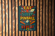 a colorful sign with the text "EAT SLEEP PINBALL REPEAT" displayed on a wooden wall.