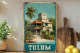 A vintage-style metal sign with a tropical house illustration, placed on a wooden shelf next to a wooden cutting board and some potted plants.