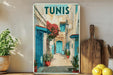 A framed poster of a colorful street scene in Tunisia, featuring blue doors, red flowers, and a cobblestone path.
