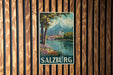 A vintage-style poster of Salzburg, Austria, featuring a scenic landscape with a mountain, trees, and a river, is displayed on a wooden wall.