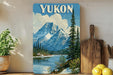 A framed vintage-style poster of a mountain landscape with a lake, pine trees, and a cloudy sky, displayed on a wall next to a wooden cutting board and some potted plants.