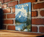 A vintage-style poster of a mountain landscape with a lake and pine trees, mounted on a brick wall.