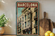 A framed vintage poster of a building in Barcelona, Spain, displayed on a shelf next to a wooden cutting board and some lemons.