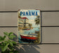A vintage-style metal sign with the word "PANAMA" prominently displayed, featuring a scenic image of boats on a river with palm trees and buildings in the background.