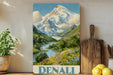 A framed painting of a mountain landscape with a river and flowers, displayed on a shelf next to a wooden cutting board and some potted plants.