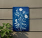 a blue sign with white flowers on it, hanging on a wall next to a plant.