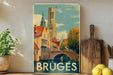 A vintage-style poster of the city of Bruges, Belgium, featuring a church with a bell tower and a bridge over a canal. The poster is displayed on a wall next to a wooden cutting board and some potted plants.