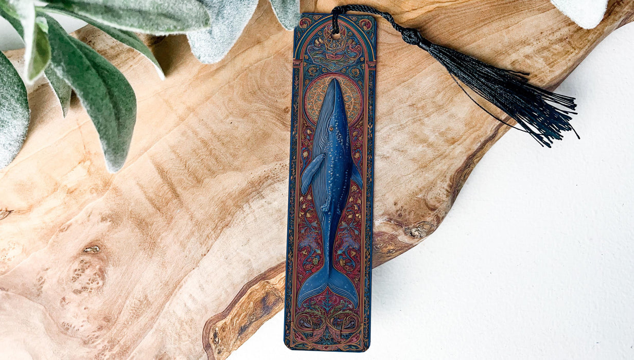 A decorative bookmark featuring a blue whale design is displayed on a wooden surface, with a black tassel hanging from the top.