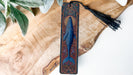 A decorative bookmark featuring a blue whale design is displayed on a wooden surface, with a black tassel hanging from the top.