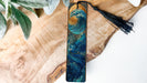 A wooden bookmark with a wave design and a black tassel is displayed on a wooden surface.