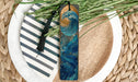 A wooden bookmark with a wave design and a black tassel is placed on a plate with a striped pattern and some green foliage.
