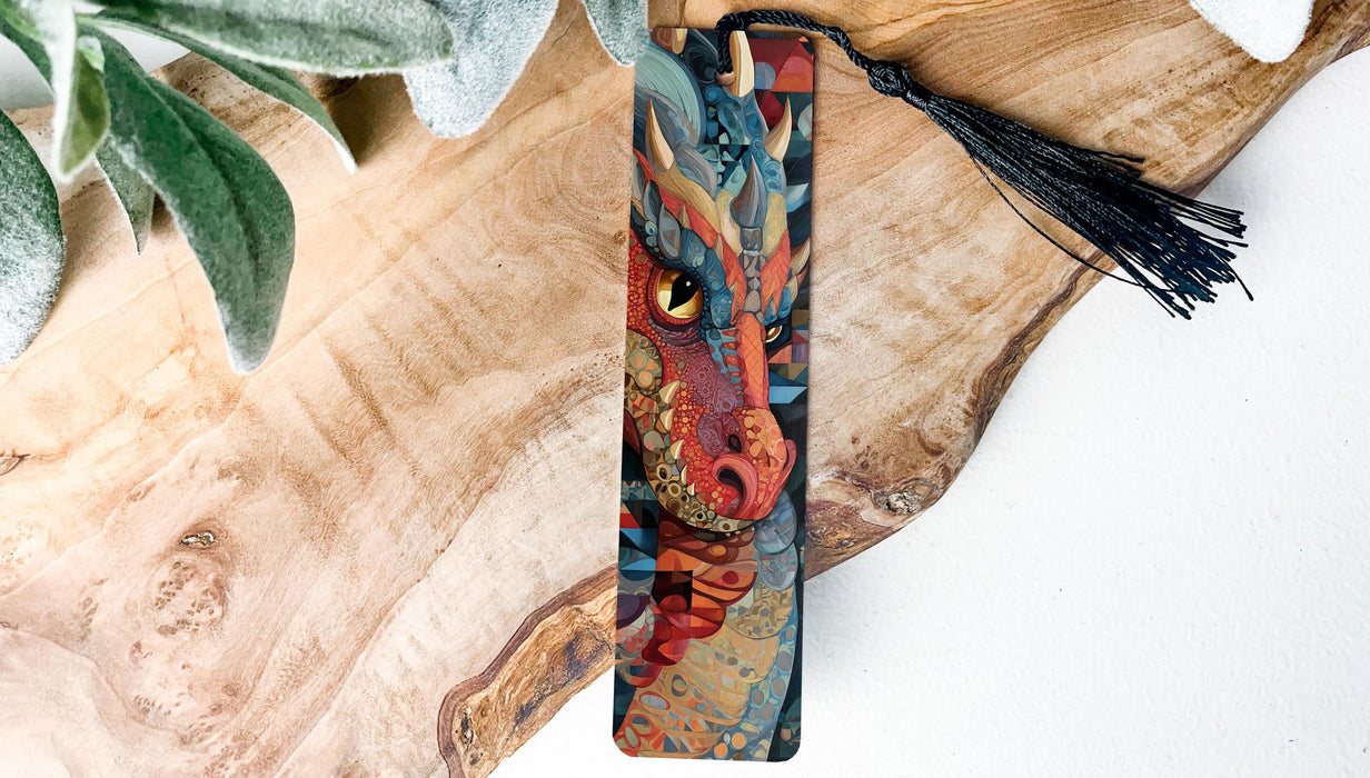 A colorful dragon-themed bookmark with a black tassel is displayed on a wooden surface, accompanied by some greenery.