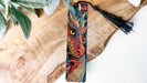 A colorful dragon-themed bookmark with a black tassel is displayed on a wooden surface, accompanied by some greenery.