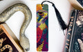 A colorful bird-like bookmark with a black tassel is placed on a white surface next to a book and a decorative snake.
