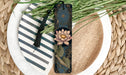 A decorative bookmark with a floral design and a tassel is placed on a plate with a striped pattern, surrounded by green grass.