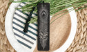 A decorative black bookmark with a feather design rests on a plate with a striped pattern, surrounded by green foliage.