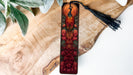 A decorative bookmark with a peacock design, featuring a vibrant red and orange color scheme with intricate floral patterns.