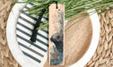 A wooden bookmark with a black tassel and a printed illustration of a beaver is placed on a plate with a woven placemat.