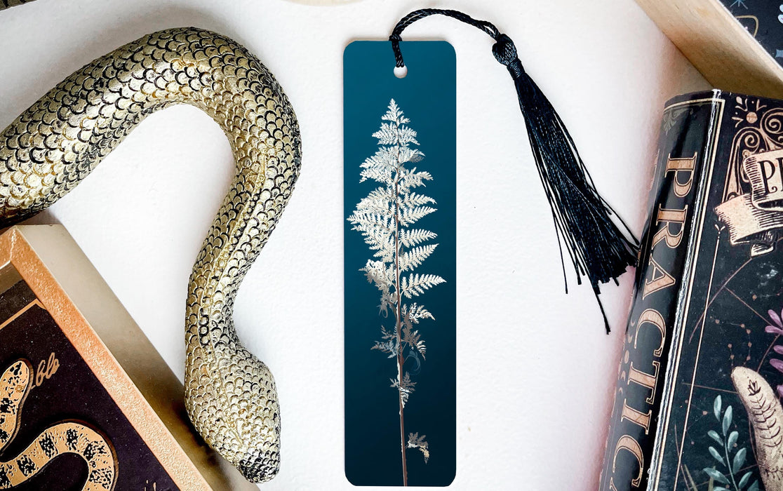 a blue bookmark with a white fern design, a black tassel, and a decorative snake figurine, all placed on a white surface.