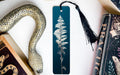 a blue bookmark with a white fern design, a black tassel, and a decorative snake figurine, all placed on a white surface.