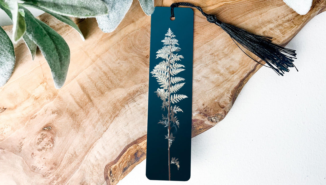 A vertical bookmark with a black tassel attached, featuring a detailed image of a fern plant on a dark blue background.