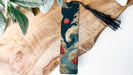 A vertical wooden bookmark with a tassel attached, featuring an illustration of a wave and a red sun in the sky.