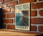 A vintage-style poster of Pulpit Rock in Norway, featuring a cliff with a group of people standing on the edge, is displayed on a brick wall.