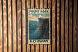 A vintage-style poster of Pulpit Rock in Norway, featuring a cliff with a river below, is displayed on a wooden wall.