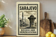 a vintage-style stamp or sign with the text "SARajevo Bosnia and Herzegovina" on it, placed on a shelf next to a potted plant and a wooden cutting board.