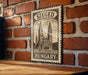 A vintage-style metal sign with the text "SZEGED HUNGARY" displayed on a brick wall.