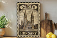 A vintage-style metal sign with a drawing of a church and the text "SZEGED HUNGARY" is displayed on a shelf, accompanied by a potted plant, a wooden cutting board, and some lemons.