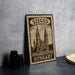 A metal sign with the text "SZEGED HUNGARY" and an illustration of a church, placed on a table next to some black camera lenses.