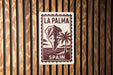 A sign with the text "LA PALMA" and "SPAIN" printed on it, along with a palm tree and a mountain, is displayed on a wooden wall.
