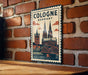 A vintage-style metal sign with a graphic of the Cologne Cathedral in Germany, mounted on a brick wall.