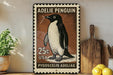 A metal sign featuring an illustration of a penguin, with the text "ADELIE PENGUIN" and "25c USA" printed on it. The sign is placed on a shelf next to a wooden cutting board and a potted plant.