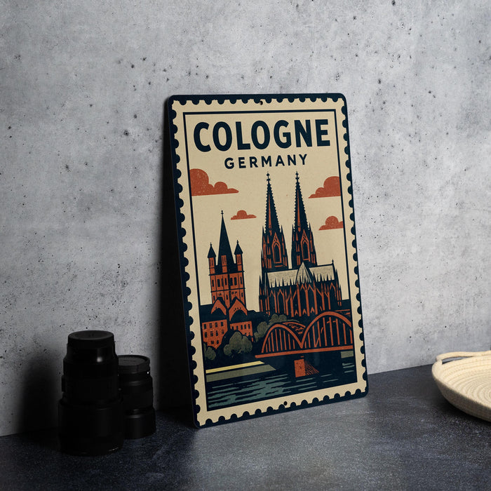 A metal sign with a vintage-style illustration of the Cologne Cathedral in Germany, featuring the cathedral's spires and surrounding buildings.