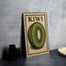 A vintage-style metal sign with a kiwi illustration, placed on a dark surface next to some black camera lenses.