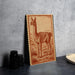 a wooden frame with a detailed illustration of a llama on it, placed on a gray countertop next to some black camera lenses.
