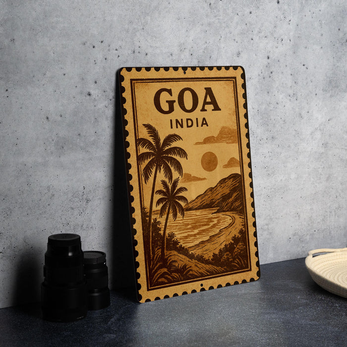 A vintage-style metal sign with the text "Goa India" and a tropical landscape illustration, placed on a gray countertop next to some black camera lenses.