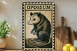 a vintage-style metal sign with a black and white illustration of an opossum, placed on a wooden surface next to a wooden cutting board and some lemons.