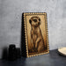 a wooden frame with a detailed illustration of a meerkat on it, placed on a dark surface next to some black camera lenses.