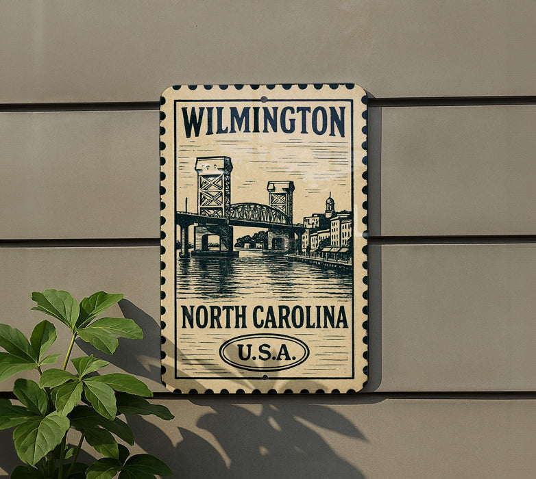 A metal sign with the text "WILMINGTON NORTH CAROLINA U.S.A." and an illustration of a bridge, mounted on a wall next to a plant.