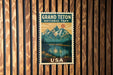 A vintage postage stamp featuring a scenic landscape with mountains, a lake, and pine trees, with the text "GRAND Teton NATIONAL PARK USA" displayed prominently.