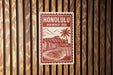 A red and white sign with the text "HONOLULU, HAWAII USA" and an illustration of a beach and mountains, mounted on a wooden wall.