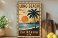 A vintage-style metal sign with the text "LONG BEACH CALIFORNIA USA" and a palm tree illustration, placed on a wooden surface next to a potted plant and a cutting board.