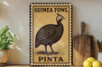 A decorative metal sign featuring a detailed illustration of a guinea fowl, with the text "GUINEA FOWL PINTA" prominently displayed. The sign is placed on a wooden surface, with a potted plant and a wooden cutting board visible in the background.