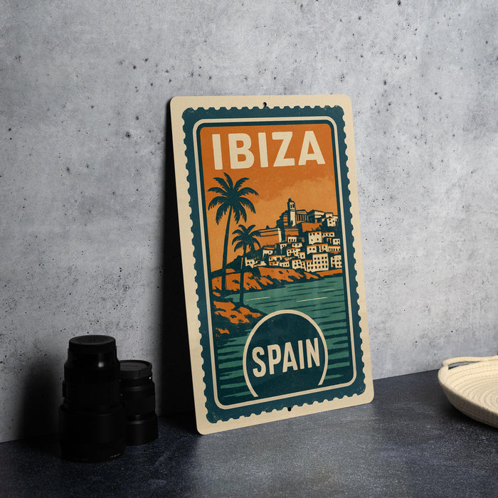 A vintage-style metal sign with the text "IBiza" and "SPAIN" printed on it, depicting a tropical island scene with palm trees and buildings.